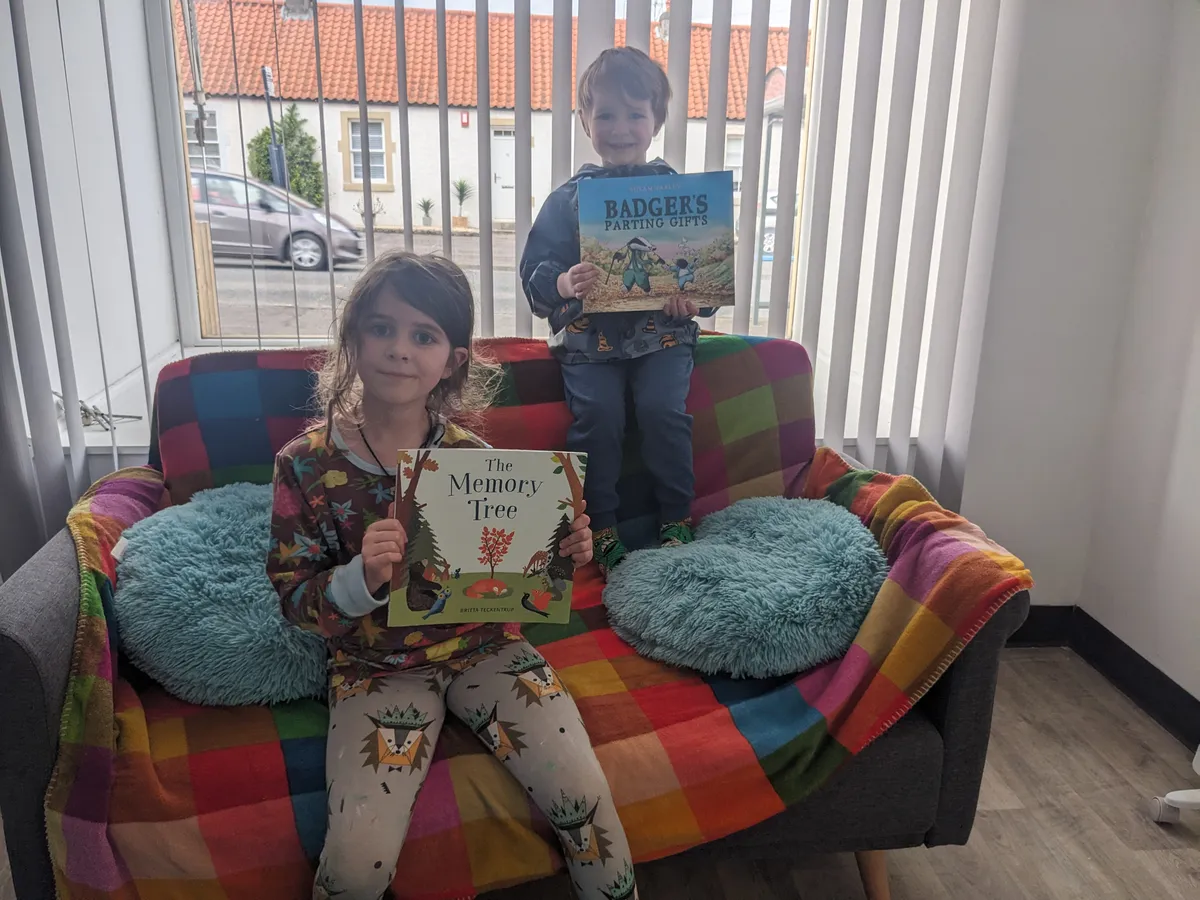 Two children on the Rainbow Room's rainbow sofa, holding the picture books The Memory Tree and Badger's Parting Gifts